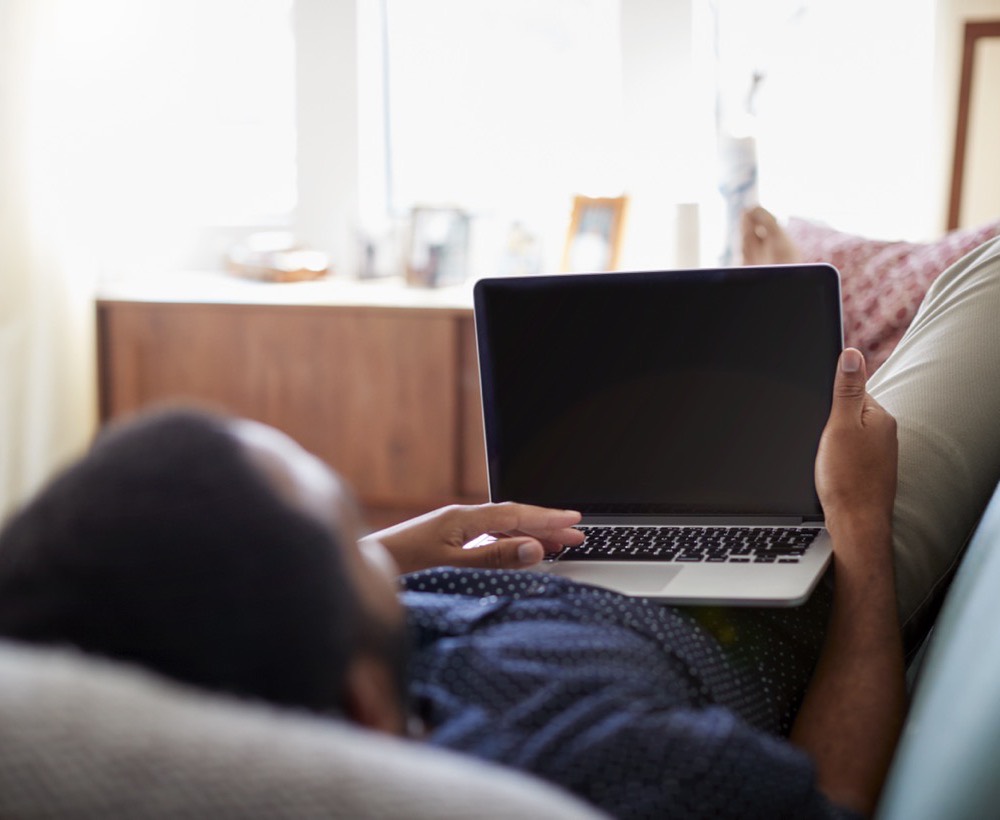 Someone lying on a sofa at home with
              a laptop resting against their knees.