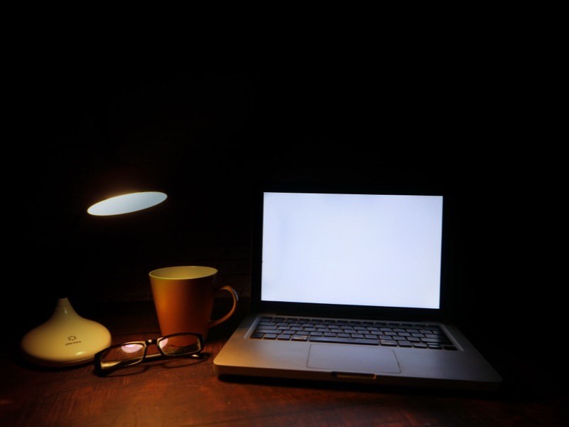 A laptop and a desk lamp on dark wood, lit in a dim room.